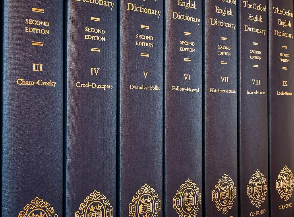 Tumpukan buku kamus tebal Oxford English Dictionary di perpustakaan universitas dengan latar belakang rak buku kayu klasik.